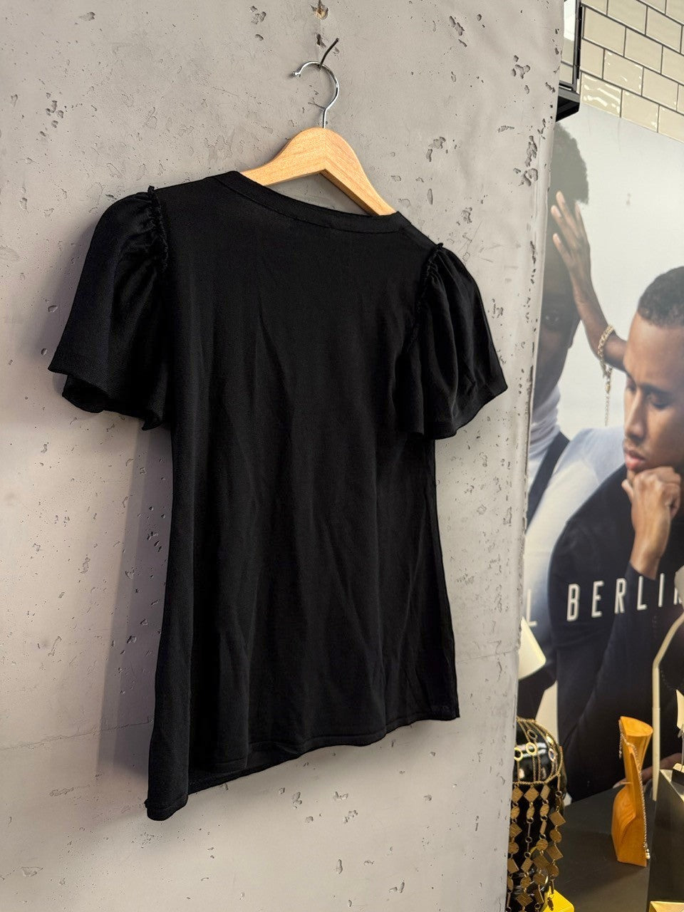 BERLIN VINTAGE CORNER black knit top with flutter sleeves hanging on a wooden hanger against a textured wall