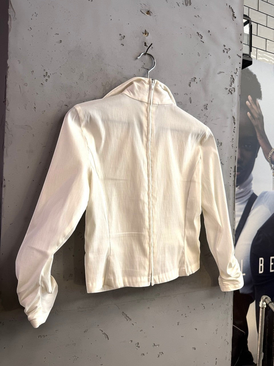 BERLIN VINTAGE CORNER cream long sleeve blouse with back zipper and ruched sleeve detail hanging on a textured wall