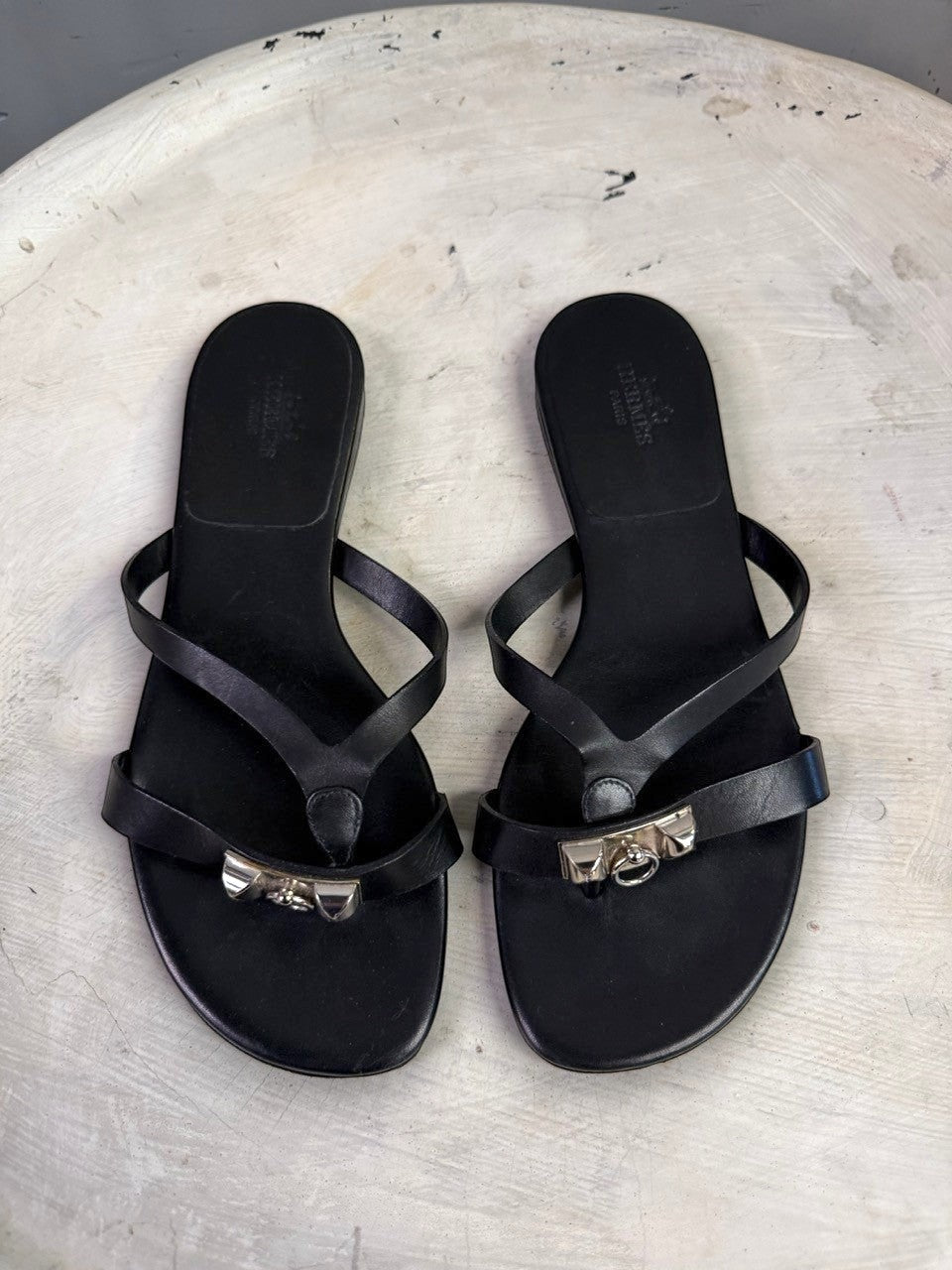 BERLIN VINTAGE CORNER black leather HERMÈS thong sandals with metallic hardware detail on a textured white surface.