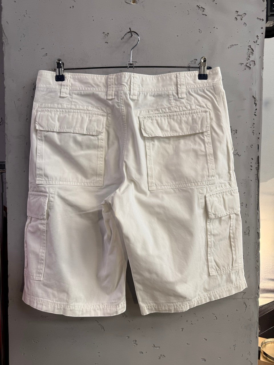 BERLIN VINTAGE CORNER McNEAL white cotton cargo shorts with multiple utility pockets, hanging against a grey wall.