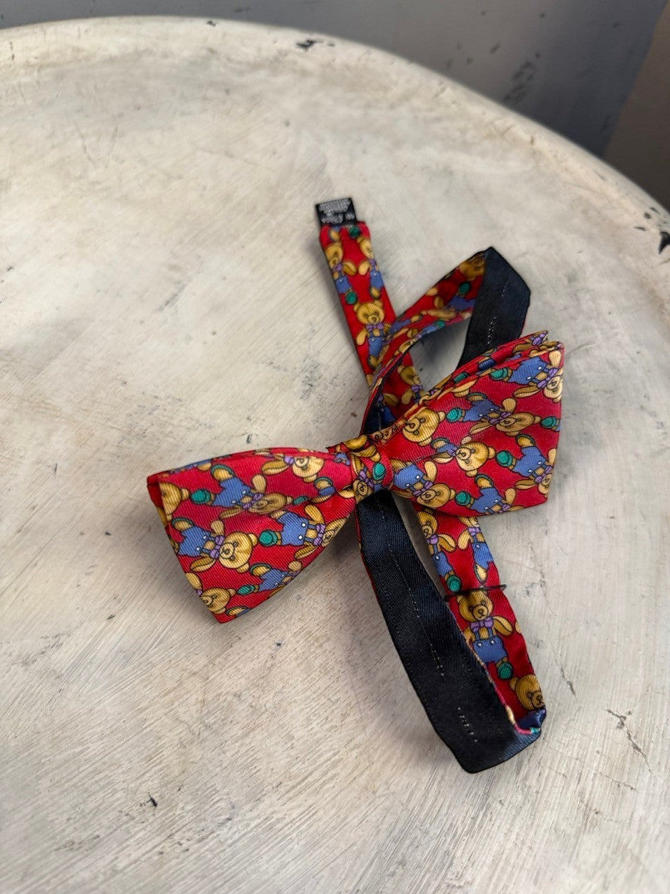 BERLIN VINTAGE CORNER red silk bow tie with teddy bear pattern. Features blue overalls and purple bows. Adjustable strap.