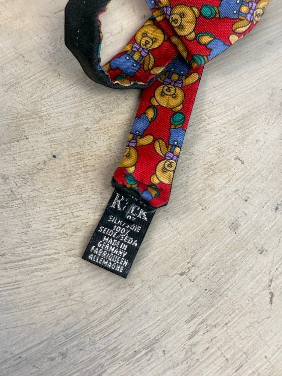BERLIN VINTAGE CORNER red 100% silk bow tie with a pattern of teddy bears in blue overalls and purple bows. Adjustable strap.