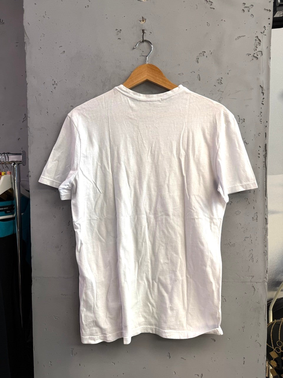 BERLIN VINTAGE CORNER white cotton crew-neck t-shirt, back view, hanging on a wooden hanger against a grey wall.